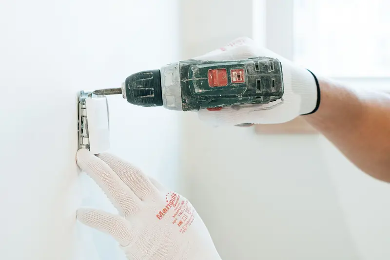 Gloved hands using a drill to install a power outlet on a wall, focusing on precision and safety